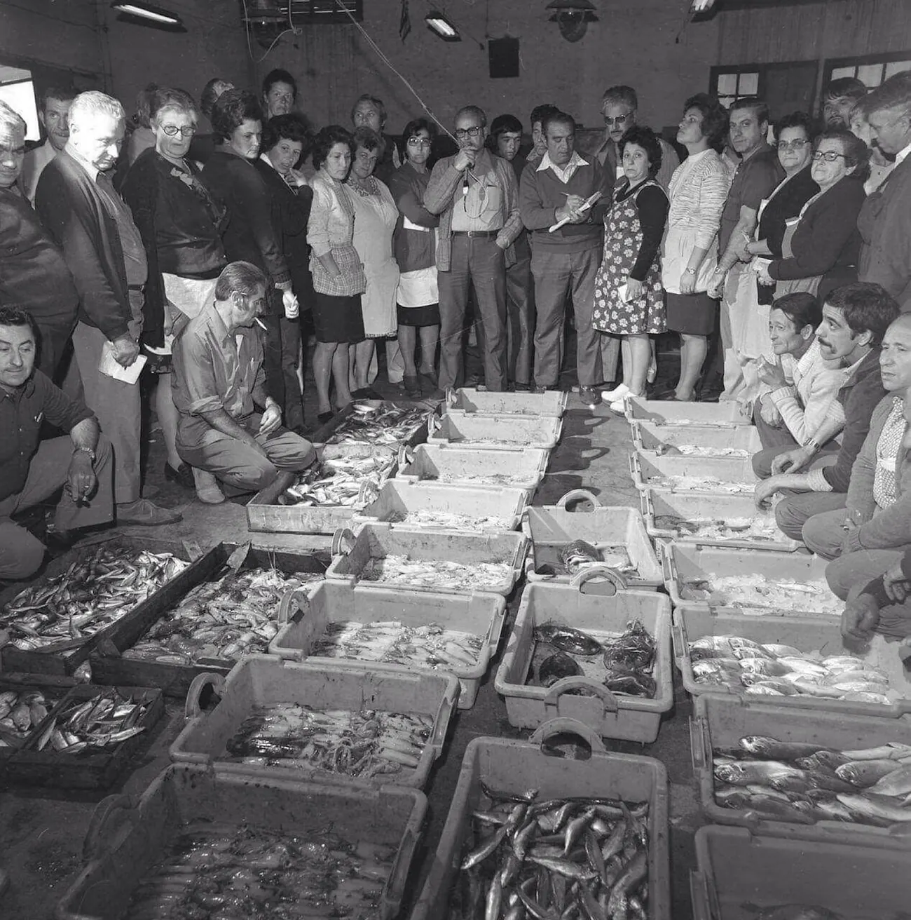 Fotografia antiga en blanc i negre de la subhasta de peix a la llotja, amb el públic al voltant de les caixes